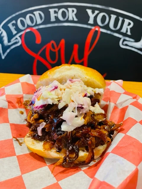 A pulled pork sandwich loaded with barbecue sauce and topped with coleslaw, served in a sandwich bun on checkered paper.