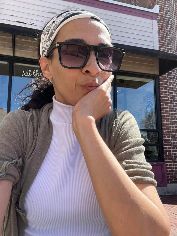 A person outdoors relaxing with sunglasses and a thoughtful expression. The background shows a brick building with windows.
