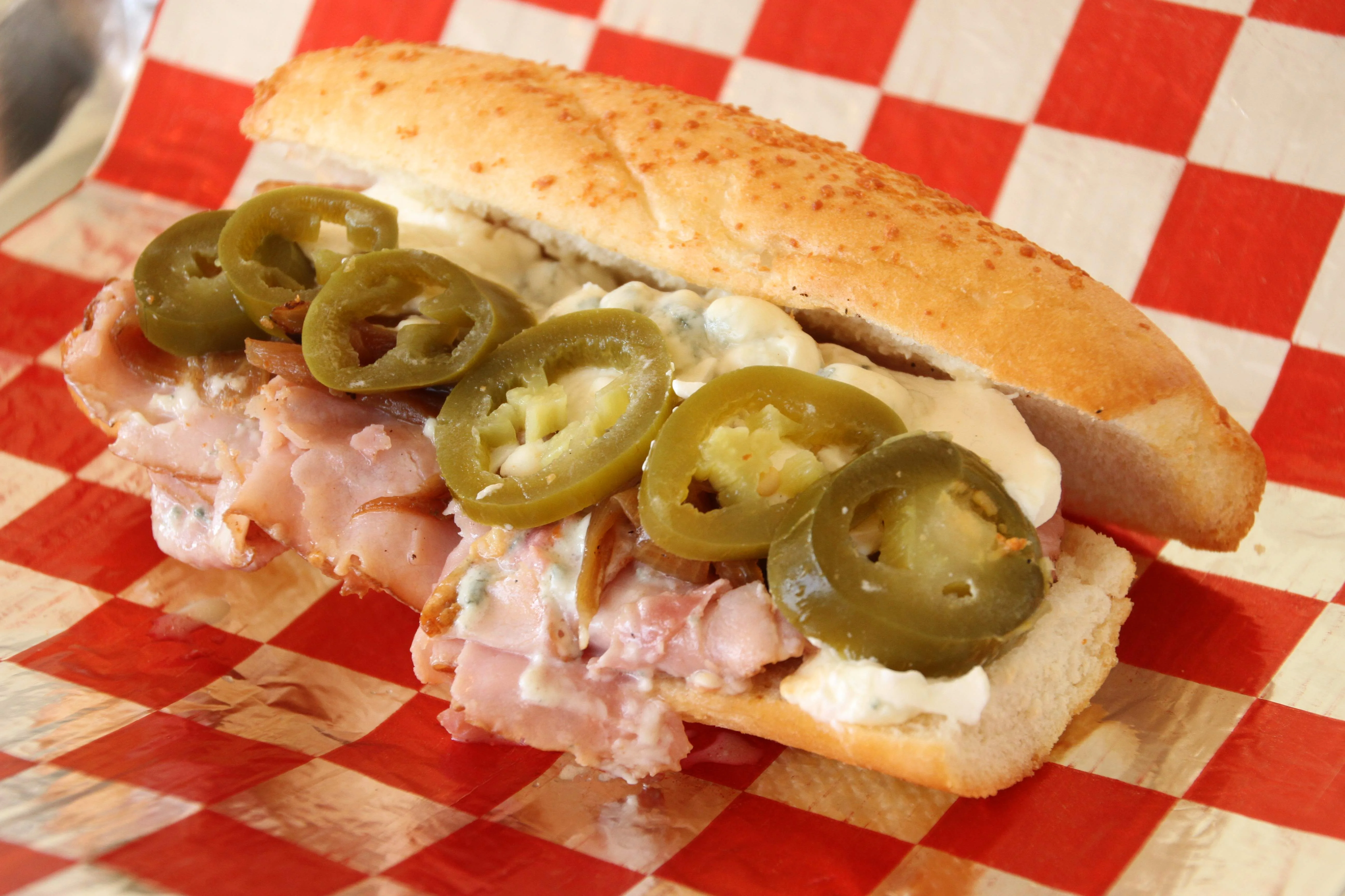 Spicy deli sandwich with sliced ham, jalapeño peppers, creamy cheese spread, and caramelized onions on a toasted hoagie roll, served on red and white checkered deli paper.