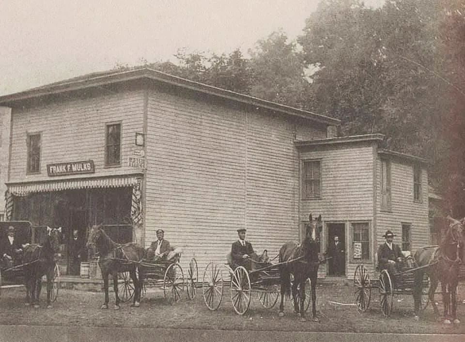 A historical black and white photo of a general store with horse-drawn carriages in front and people gathered outside.