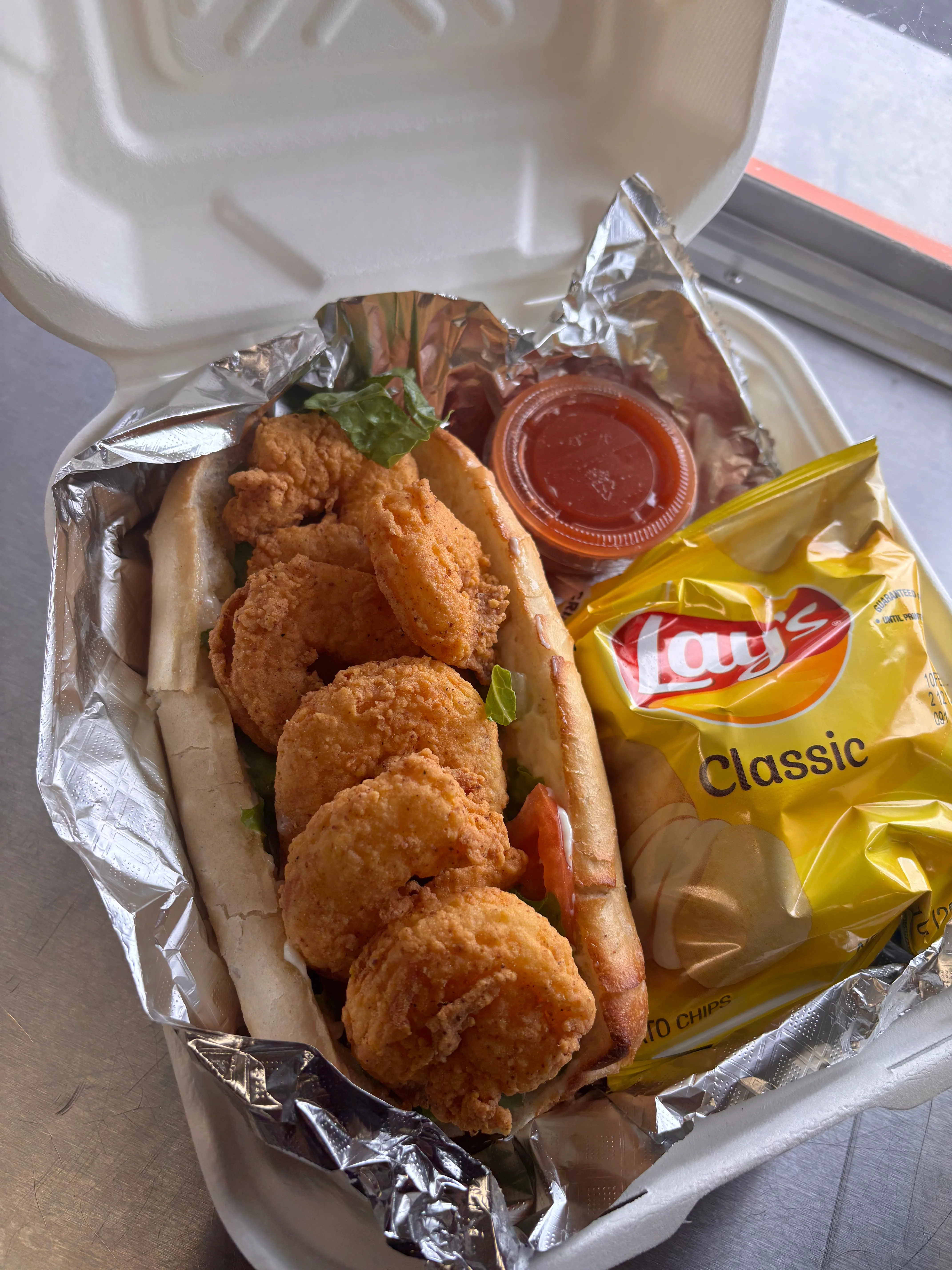 A fried shrimp sandwich served in a bun with lettuce and a side of chips and dipping sauce.