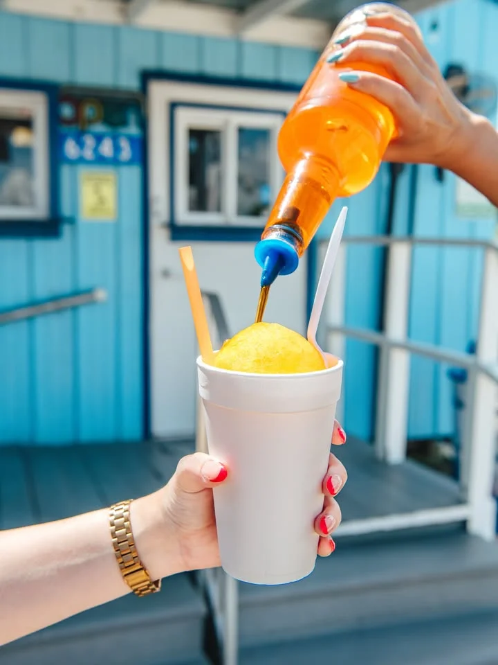 A hand pouring a bright orange syrup over a yellow shaved ice in a cup, held up against a colorful background.