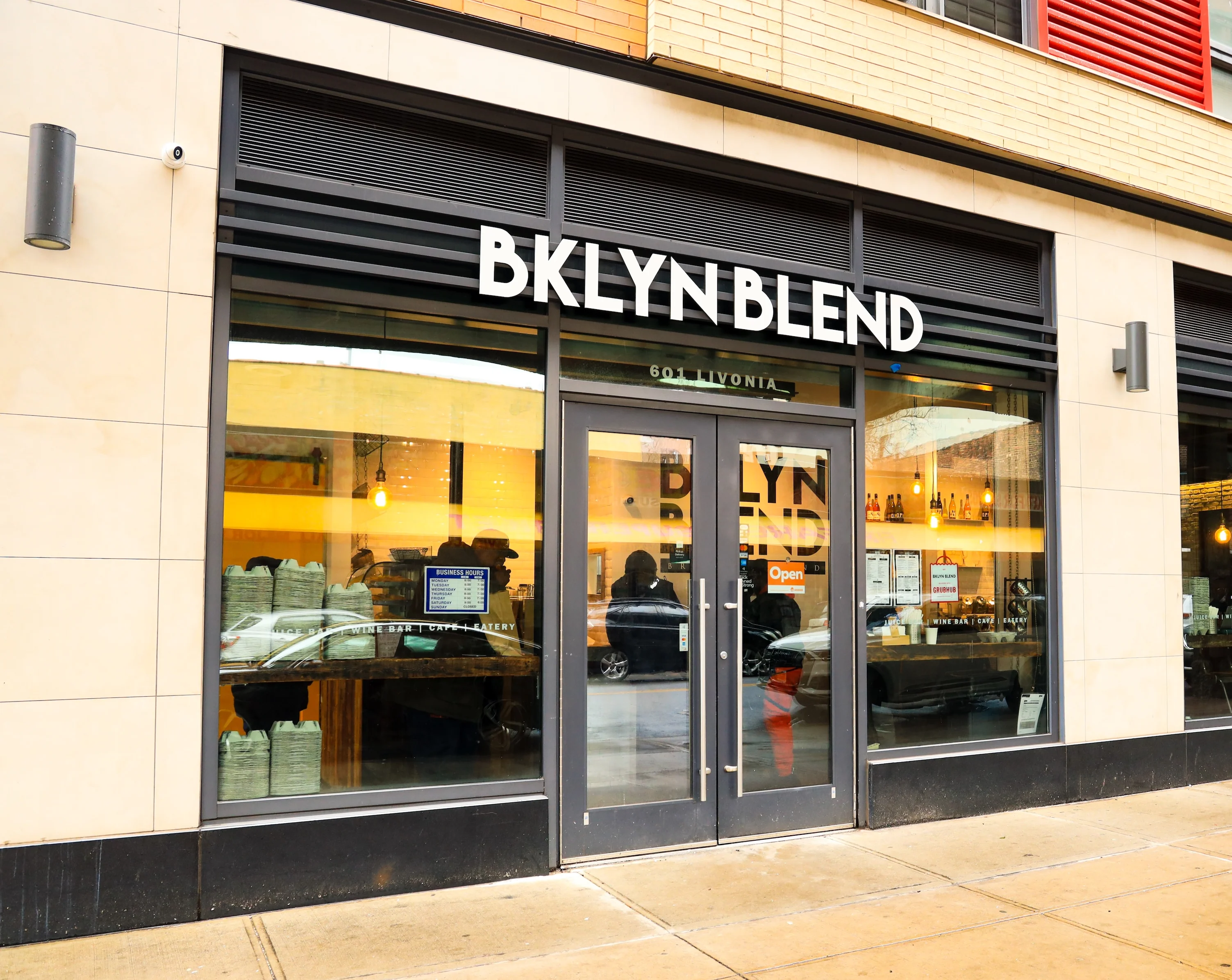 Front of building with BKLYN BLEND in large white letters above door, double glass doors with large glass windows, black door and window frames, people are inside, yellow hanging lights light up the interior