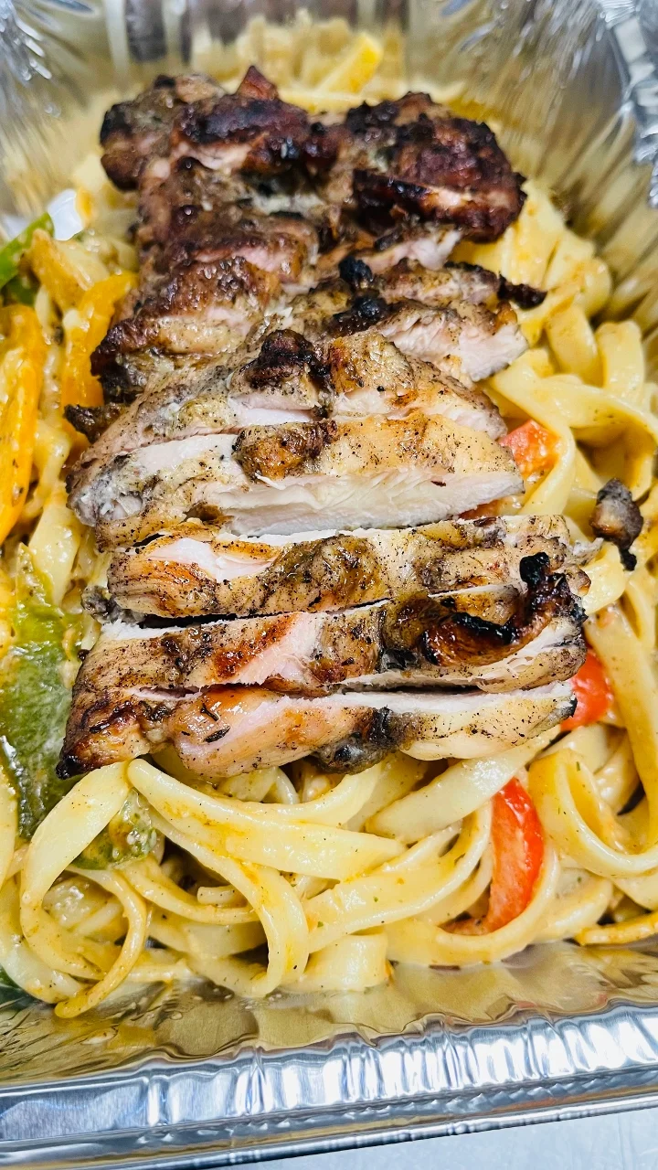 Grilled chicken slices served over a bed of fettuccine pasta mixed with colorful vegetables in a shiny takeout container.