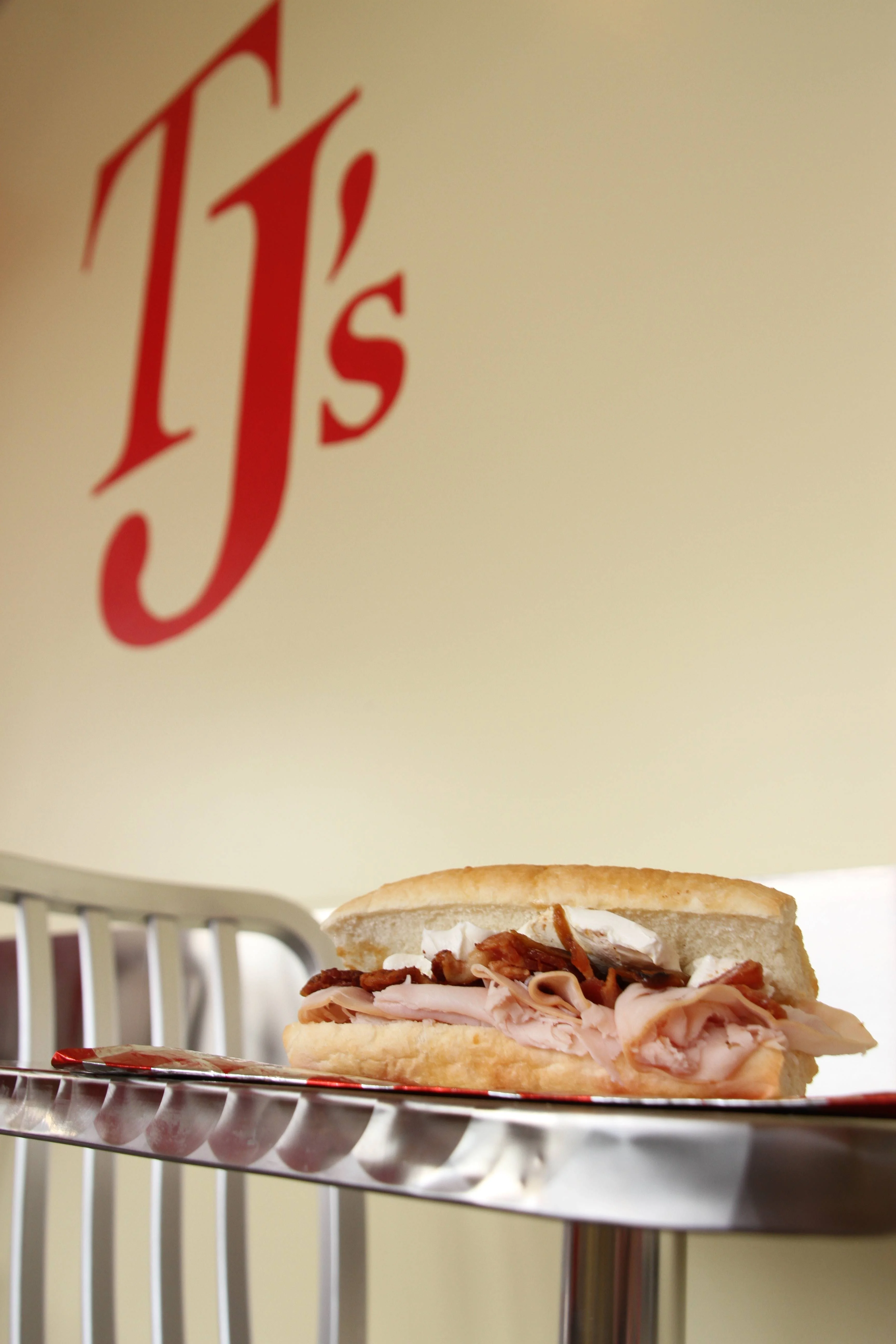A toasted sandwich filled with sliced turkey, crispy bacon, and cheese sits on a metal counter inside TJ's Sandwich Shop, with the red 'TJ's' logo visible on the wall in the background.