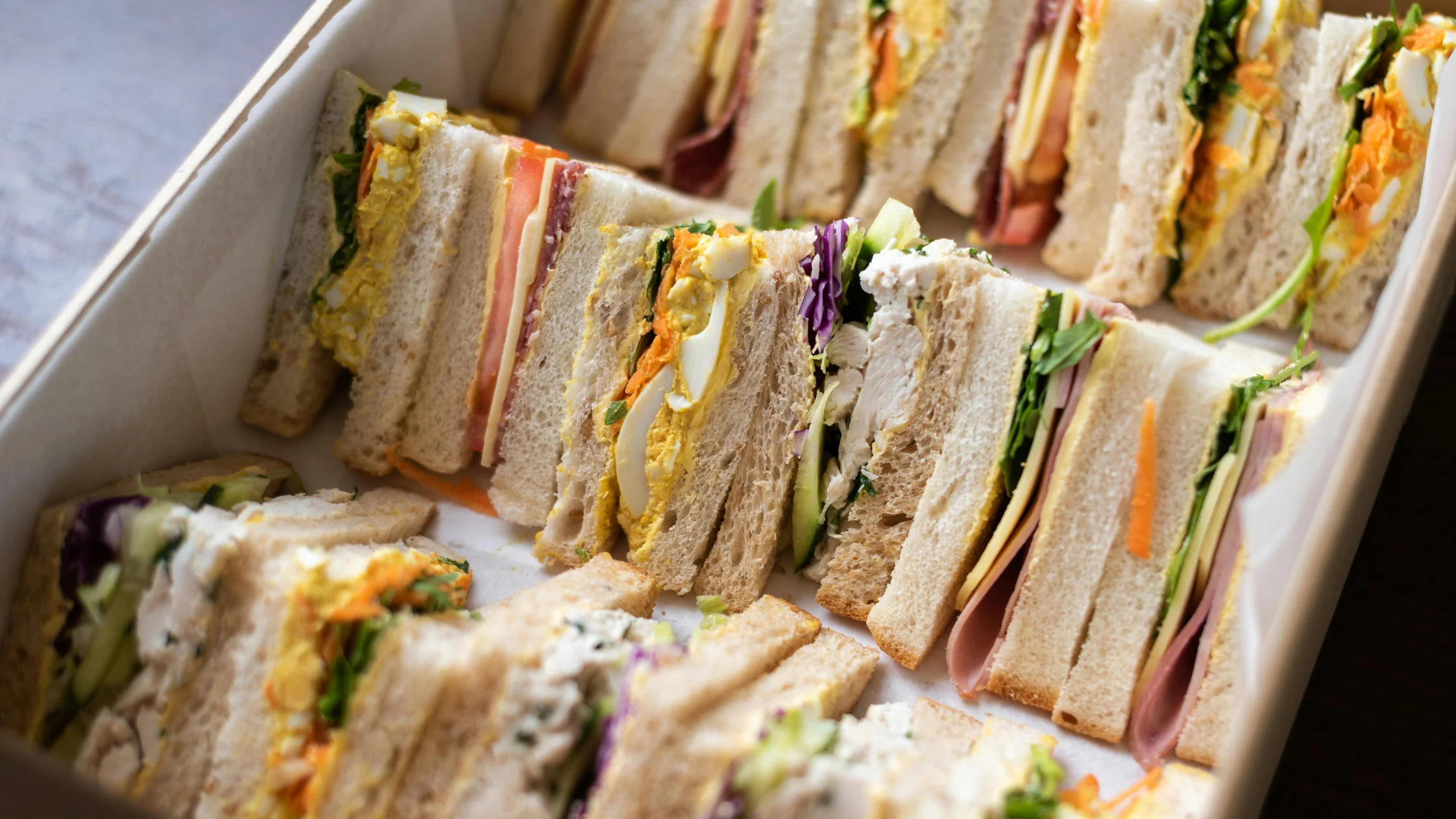 A box filled with assorted sandwich halves, neatly arranged in rows. The sandwiches are made with soft white or whole wheat bread and filled with a colorful variety of ingredients like lettuce, cheese, ham, shredded carrots, egg, and sliced vegetables.