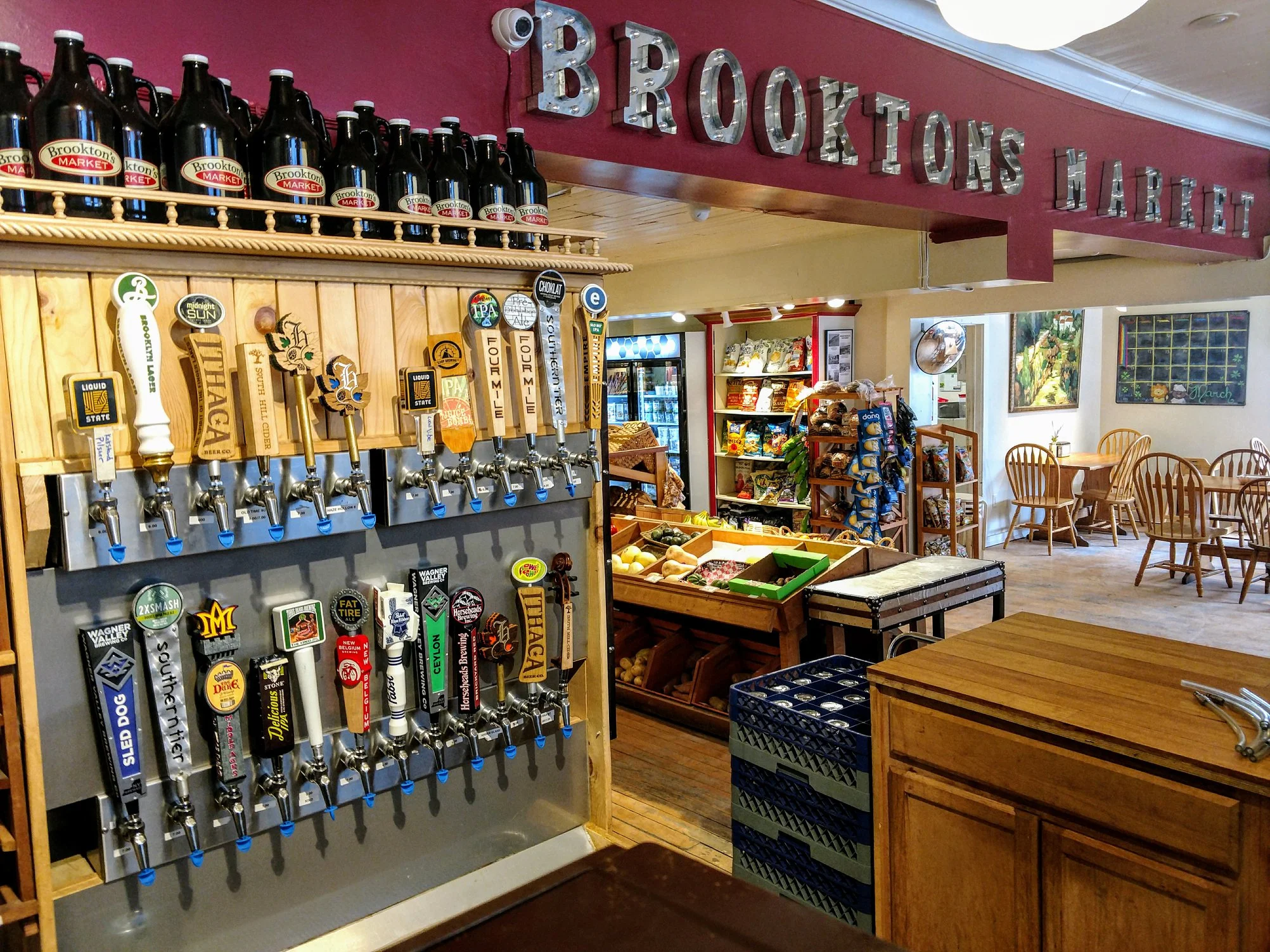 A bar setup with various beer taps on display, bottles lined up above, and a market area in the background.