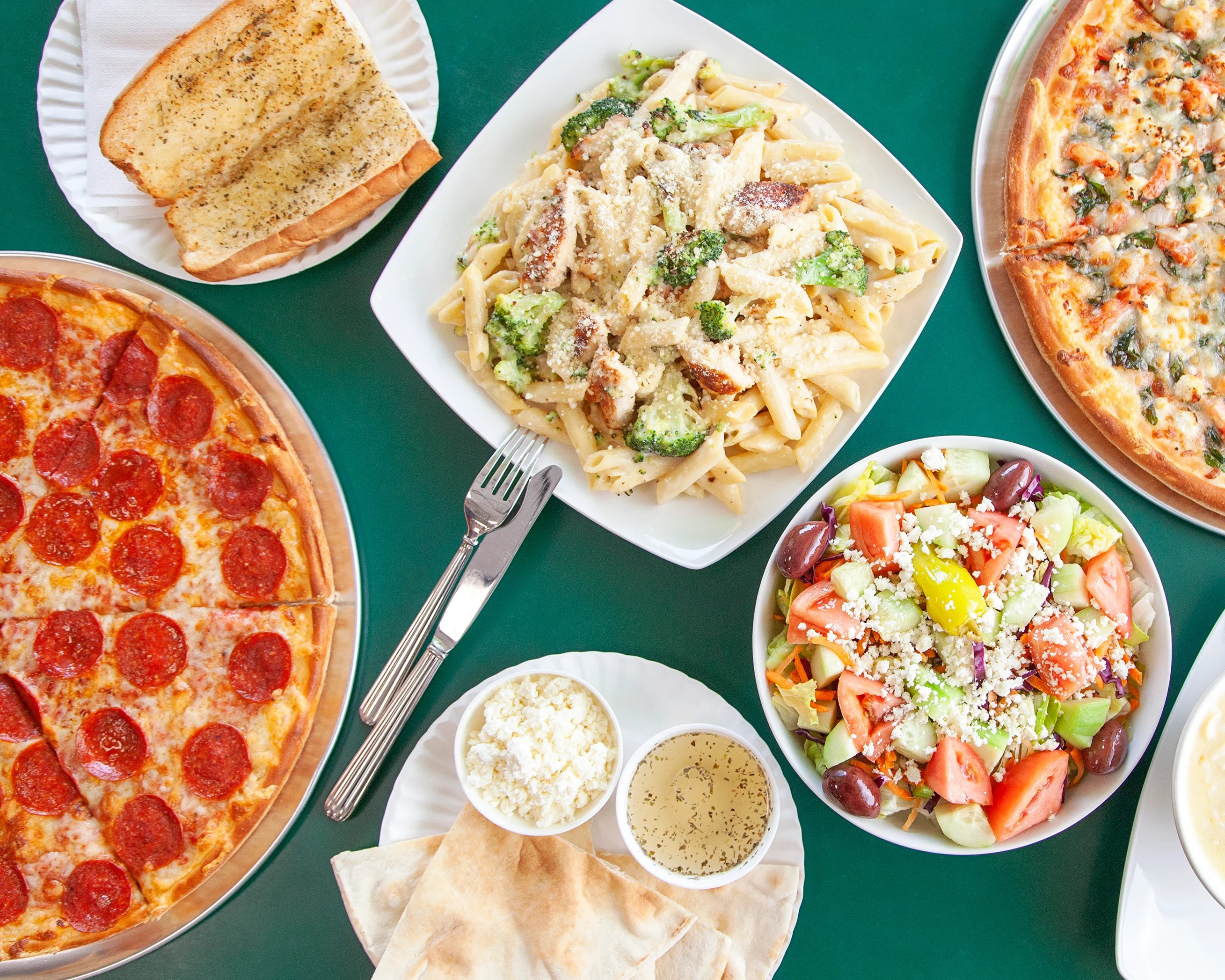 Overhead spread of pepperoni pizza, creamy pasta, Greek salad, and garlic bread