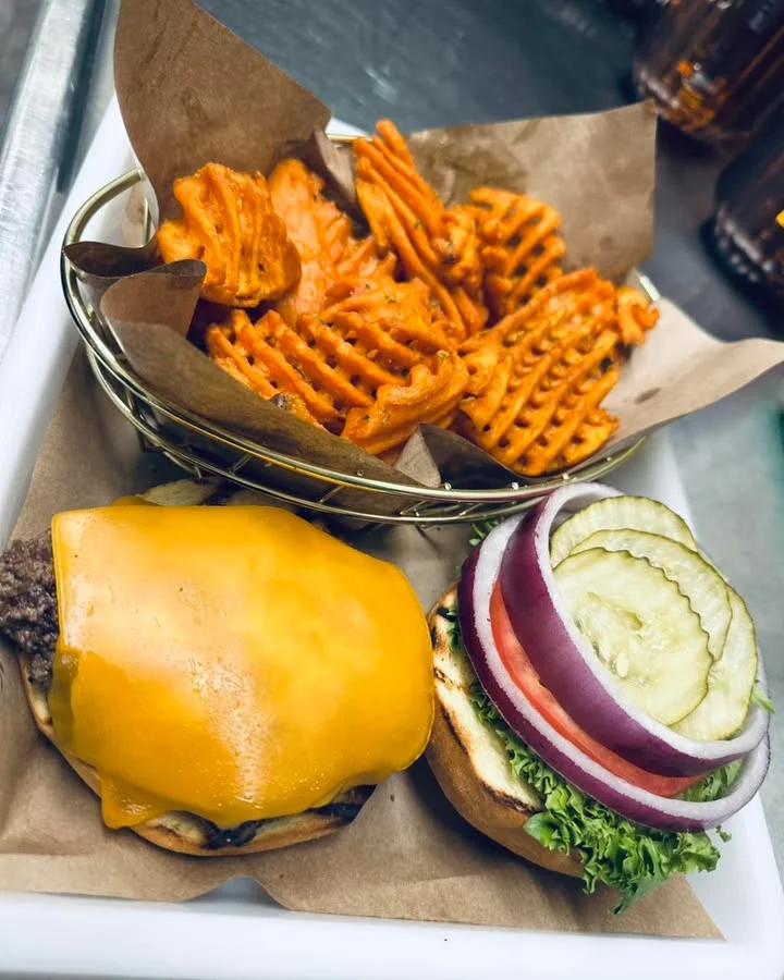 A delicious meal featuring a cheeseburger with a slice of cheddar cheese, fresh vegetables, and a side of sweet potato fries served in a basket.