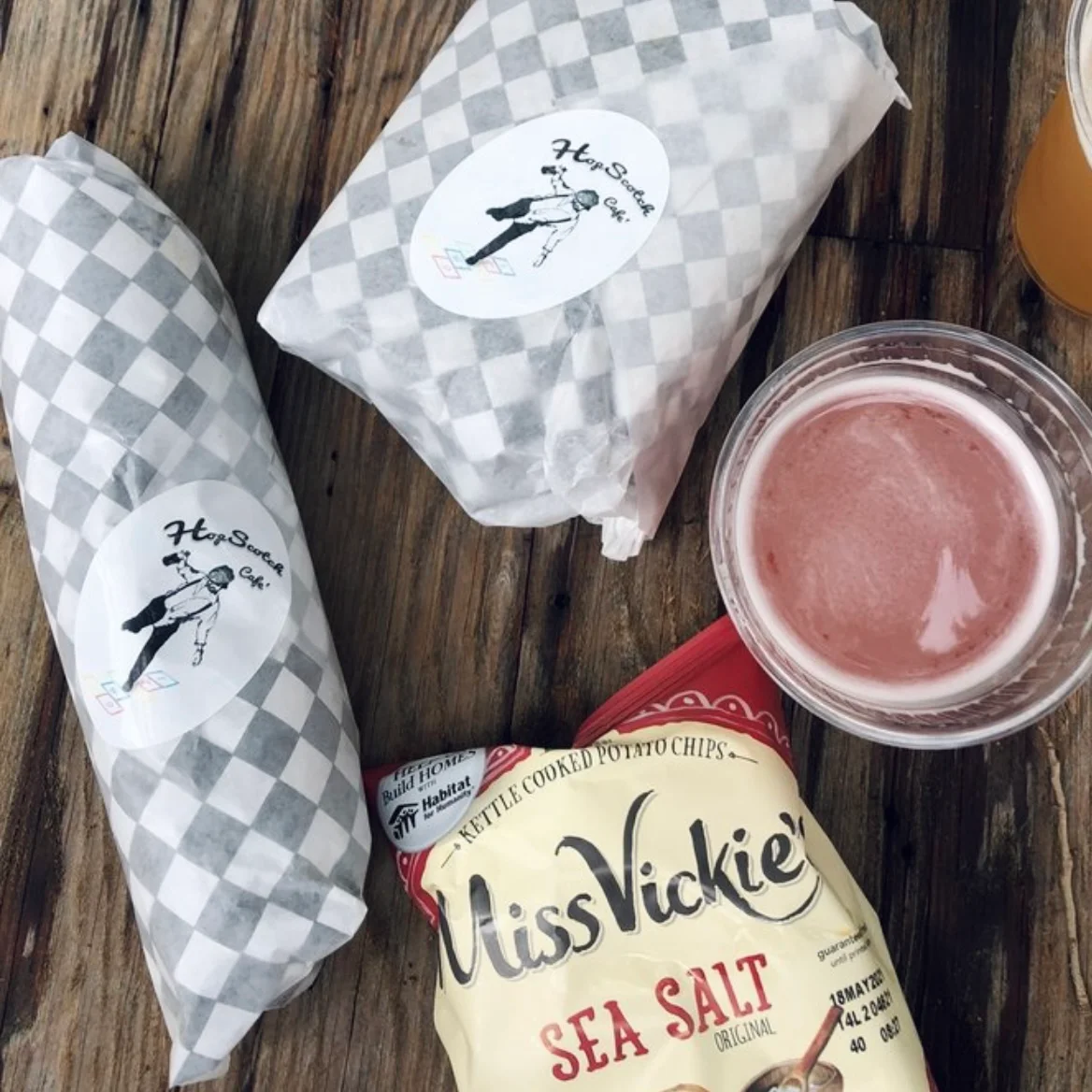 Two wrapped sandwiches with a logo, accompanied by a bag of potato chips and a cup of a pink beverage.