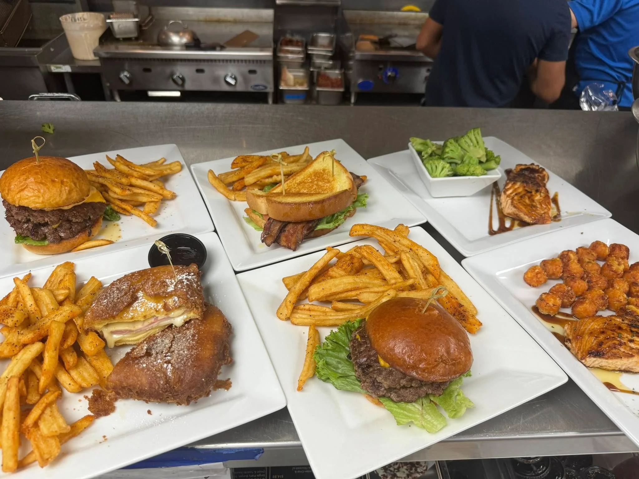 An assortment of plates including burgers with fries, grilled salmon with broccoli, tater tots, and a Monte Cristo sandwich, ready to be served.