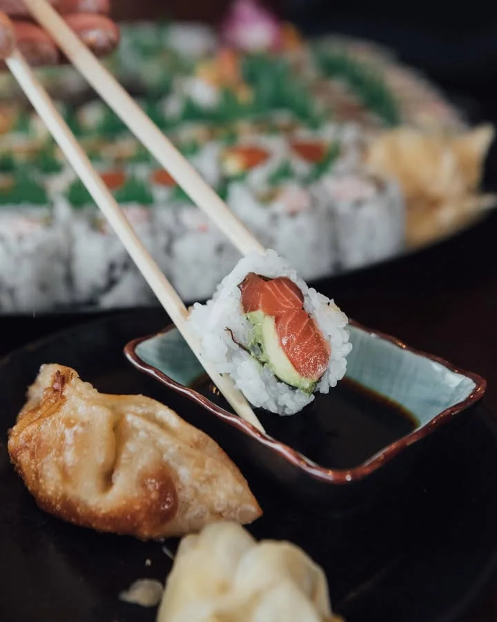 A piece of sushi held with chopsticks, accompanied by dipping sauce and a gyoza on a black plate.