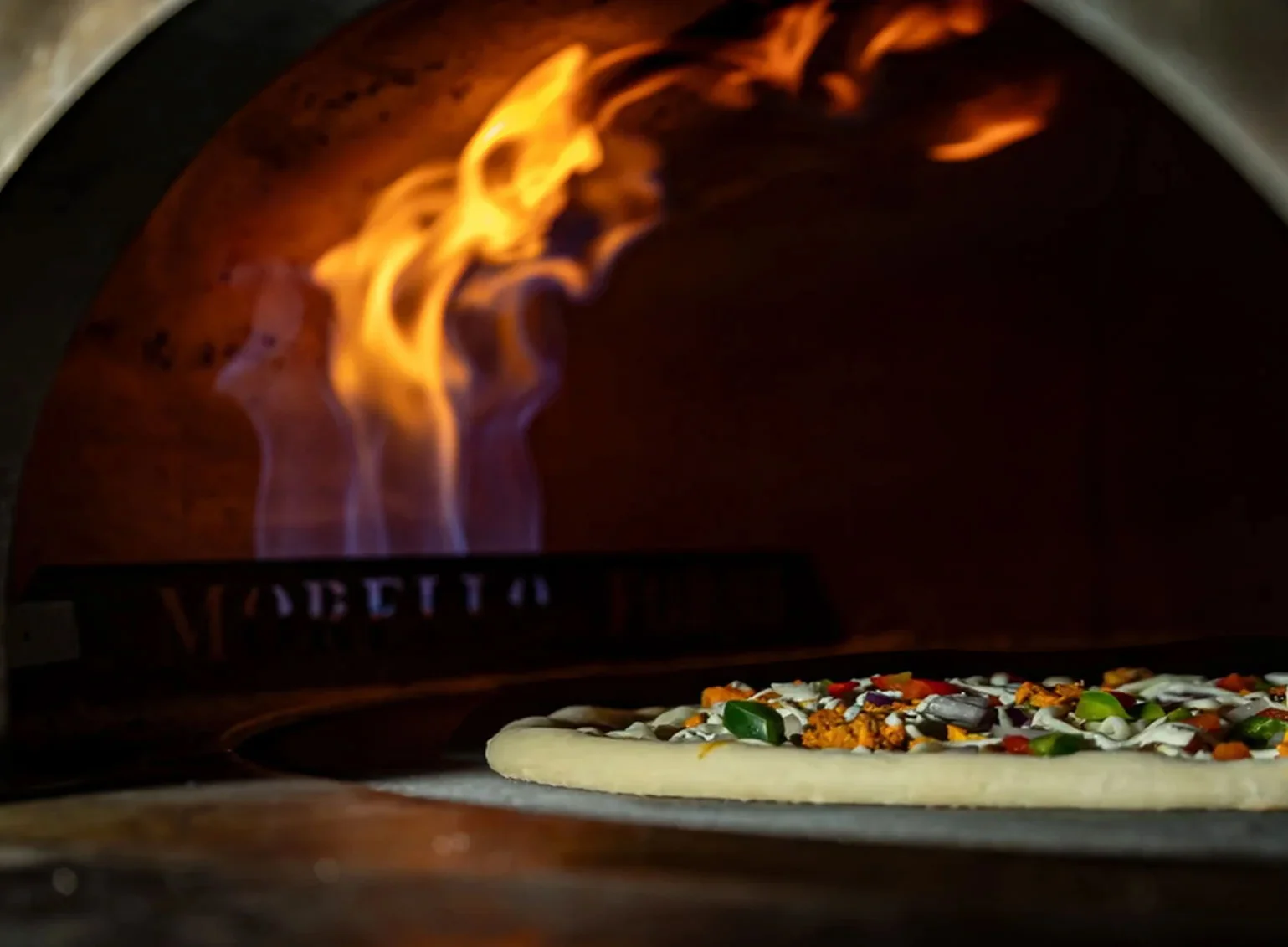 Brick-oven pizza baking inside Noosh Nosh’s artisan oven in Louisville, KY, featuring fresh toppings like green peppers, red onions, and seasoned meat, with visible flames and the “MORELLO” oven brand adding rustic ambiance—highlighting local flavor and handcrafted cooking in Clifton neighborhood.