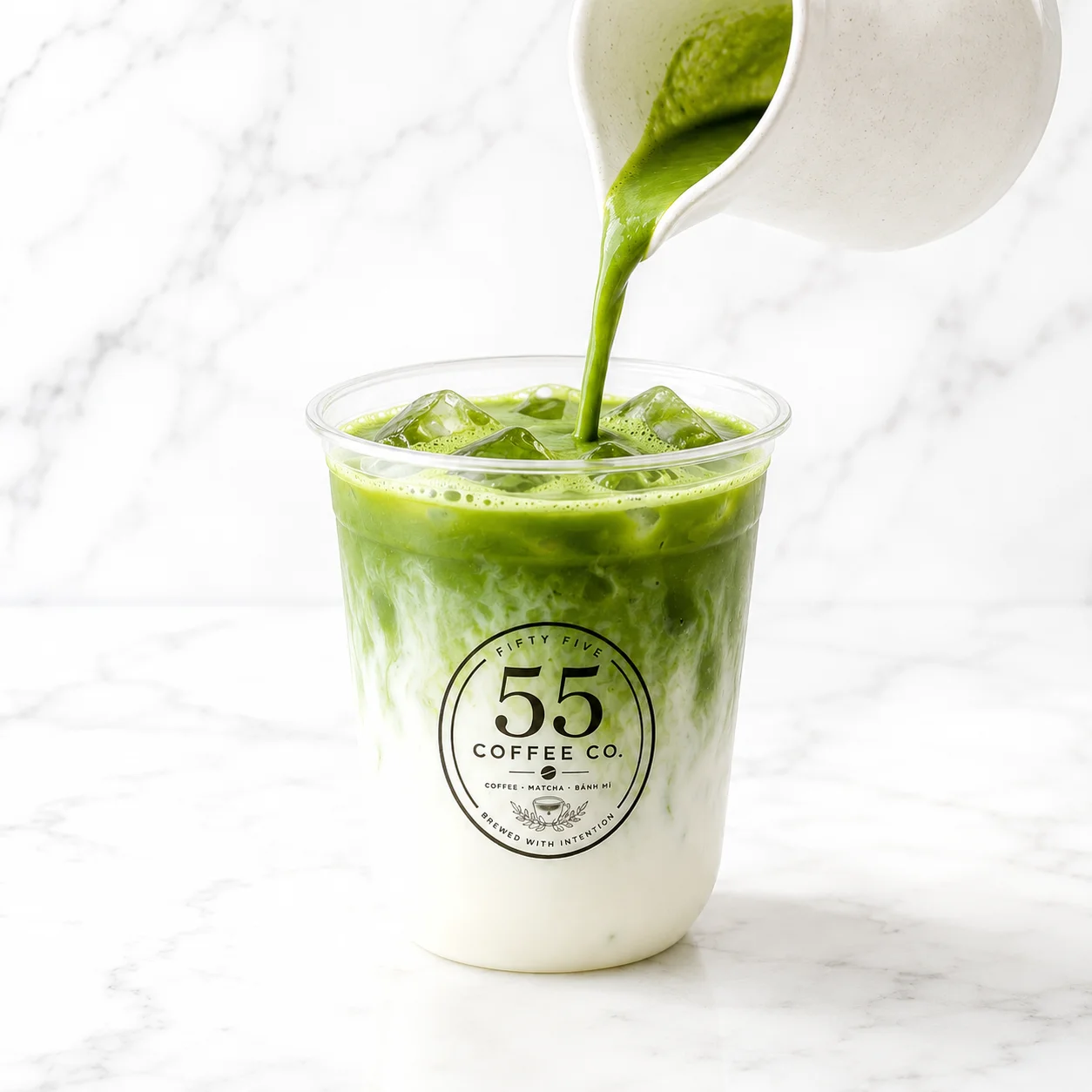 A cup filled with iced green matcha, layered with milk, showing a vibrant green color topped with frothy milk. The cup is branded with a logo.