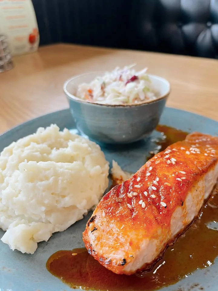A serving of salmon fillet with a sweet glaze, accompanied by fluffy mashed potatoes and a side salad in a bowl, beautifully arranged on a plate.