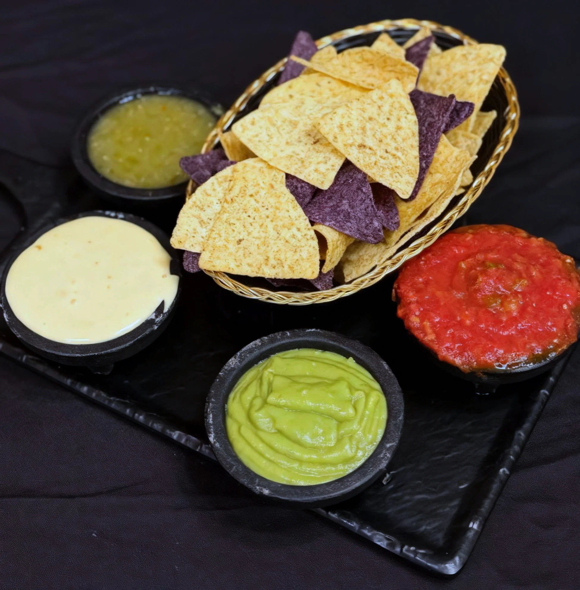 A basket of colorful tortilla chips accompanied by four bowls of various dips including cheese sauce, green sauce, and salsa.