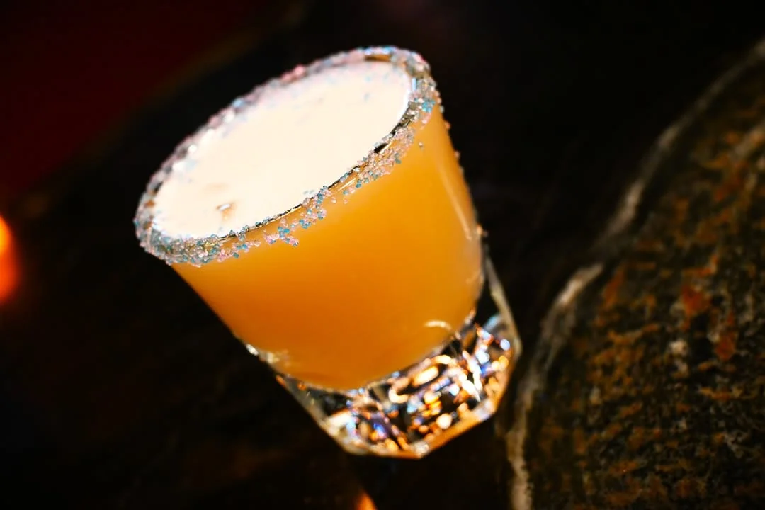 A decorative cocktail glass filled with a creamy orange drink, rimmed with sparkling sugar, sitting on a dark surface.