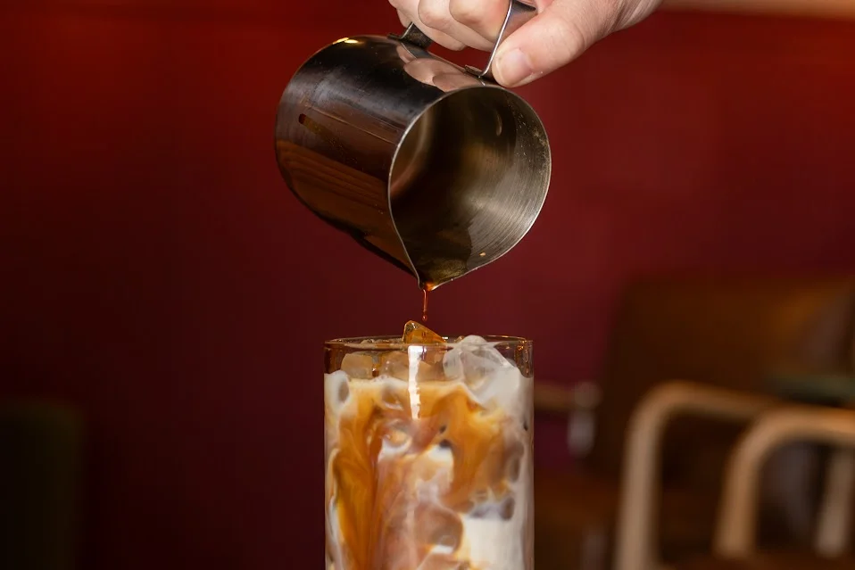 A hand pouring syrup into an iced coffee drink, creating a visually appealing mixture with swirls of color.