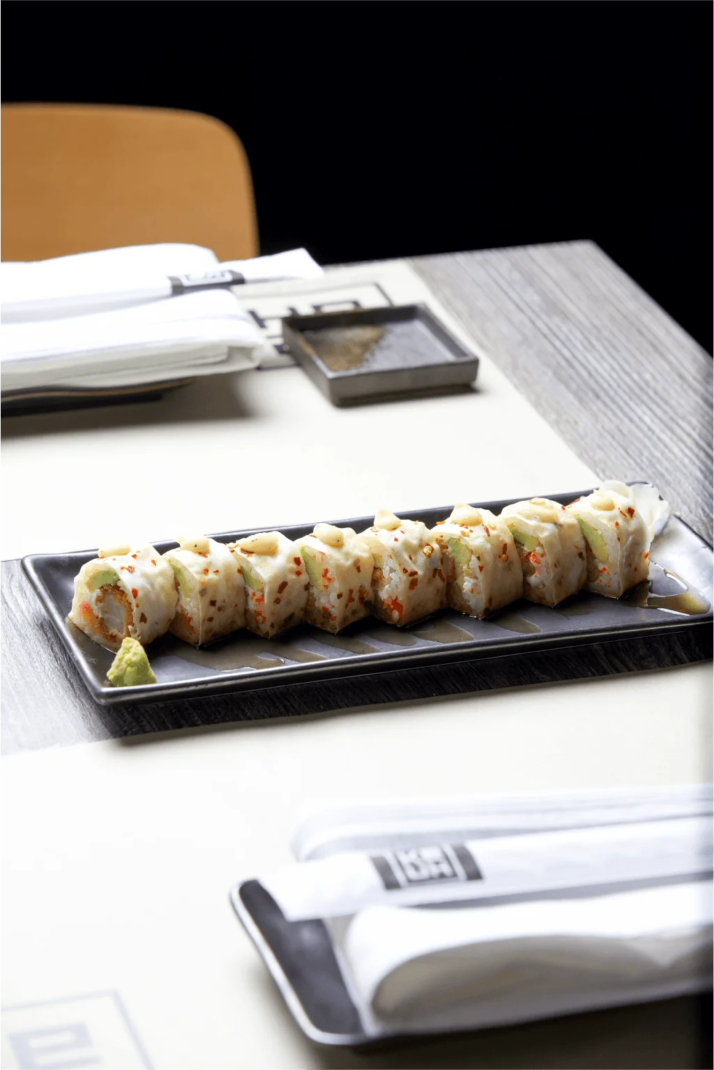 An elegant sushi roll wrapped in a white soy paper or translucent layer, sliced into eight pieces and topped with small dollops of sauce, arranged on a long dark plate with a touch of wasabi on a restaurant table.