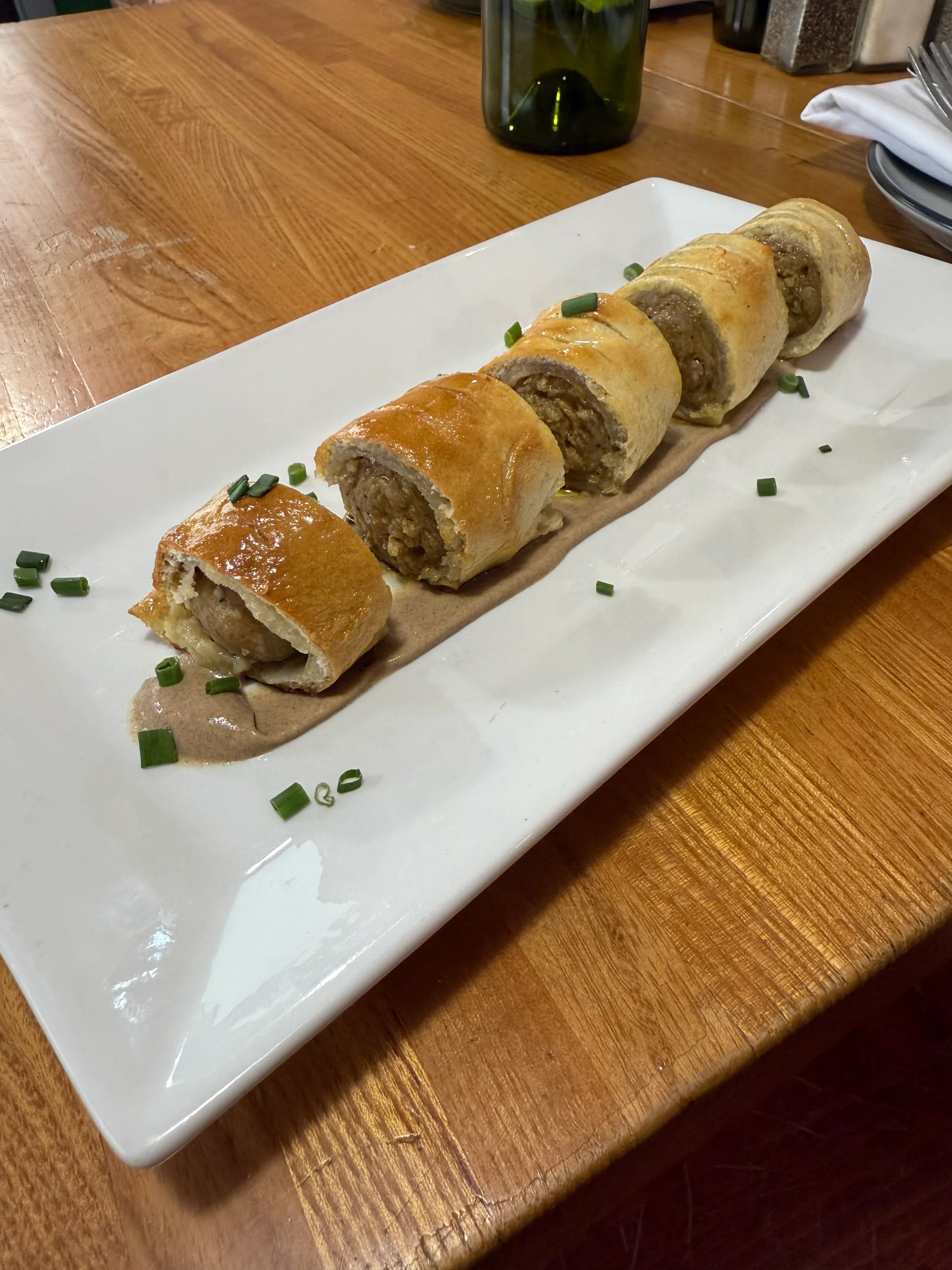 A dish featuring rolled pastry filled with meat, served on a white plate with a savory sauce and garnished with green onions.