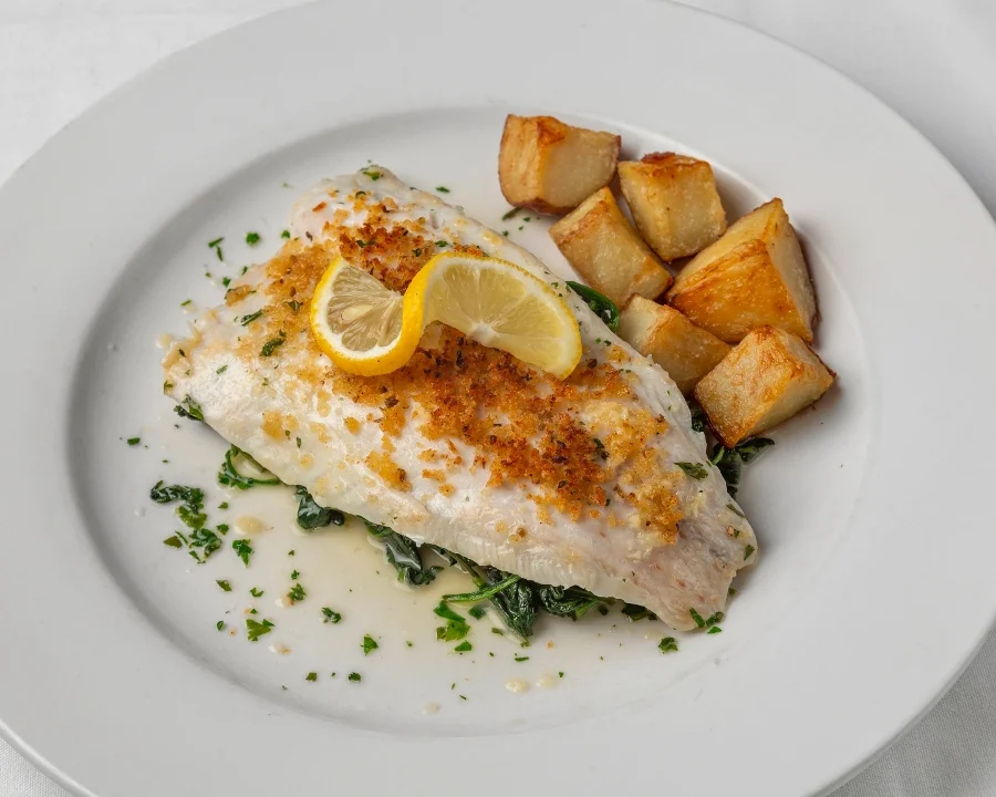 Delicious baked fish fillet topped with lemon and herbs, served with crispy roasted potatoes and fresh greens on a white plate.