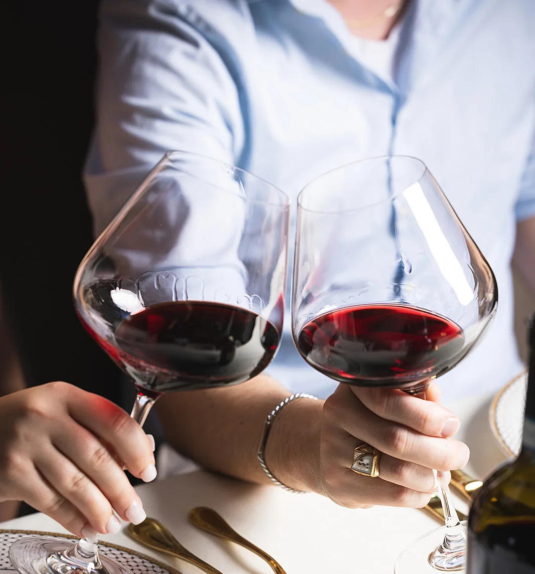 Two glasses of red wine are raised in a toast, accompanied by elegant tableware and a wine bottle in the background.