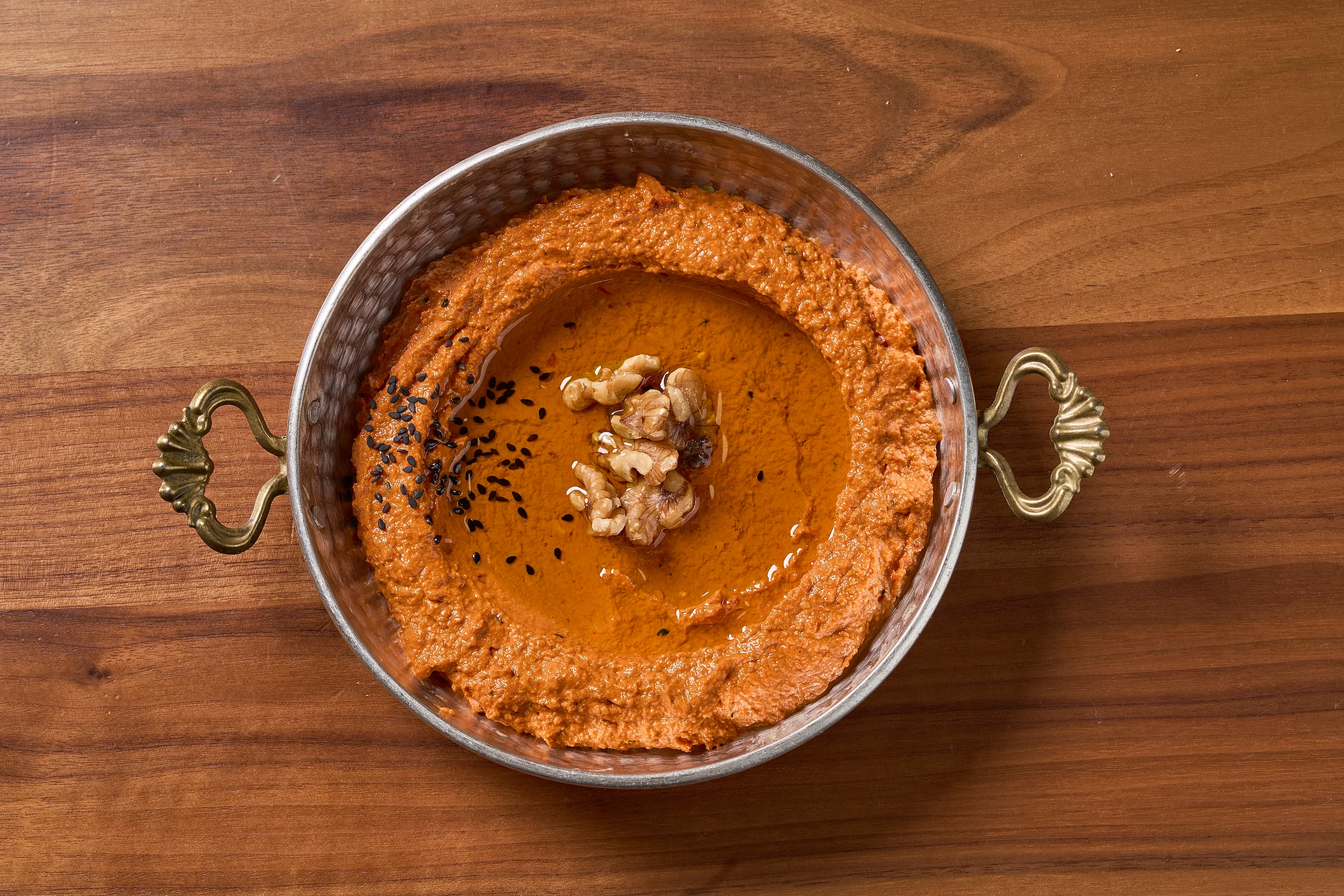A round metal bowl with handles contains smooth, reddish dip garnished with black sesame seeds and walnuts, set on a wooden surface, conveying a rustic, appetizing look.