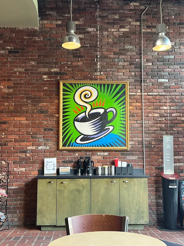 A vibrant painting of a steaming coffee cup hangs on a brick wall above a coffee station with dispensers and supplies.
