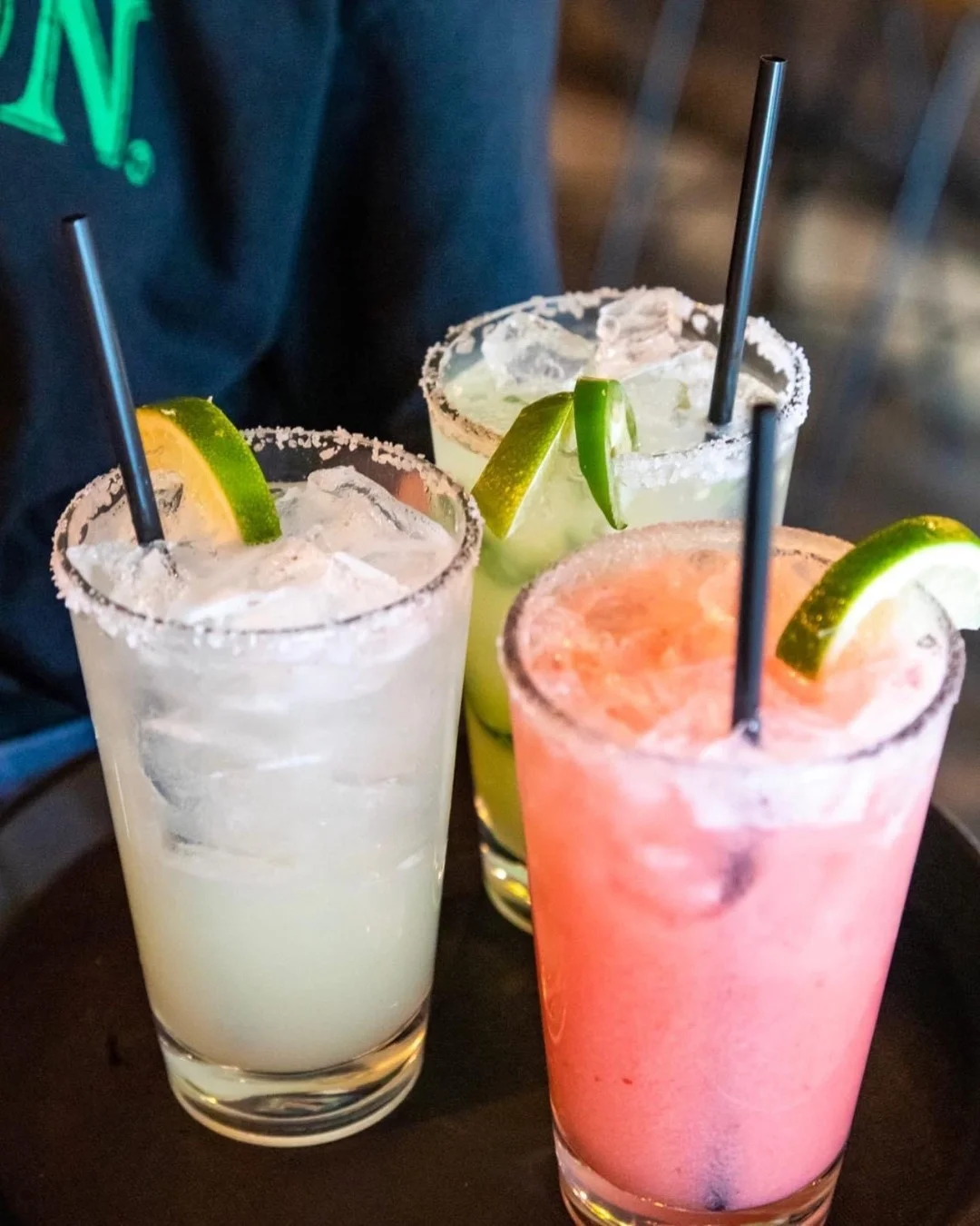 Three colorful cocktails on a serving tray, each garnished with lime slices and served with straws. Two are clear and greenish, while one is pink.