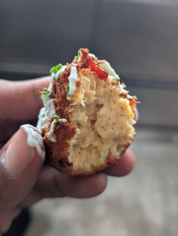 A close-up of a fried snack with a crunchy exterior and visible fillings. It is garnished with green onions and a drizzle of sauce.