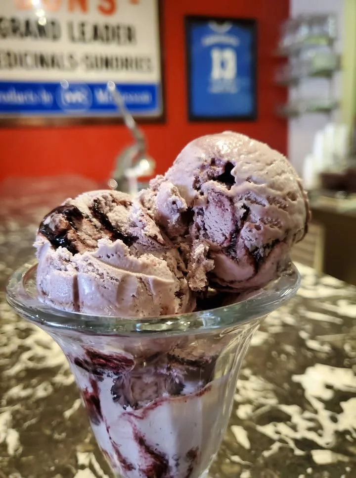 A glass dish filled with two scoops of purple-hued ice cream, swirled with chocolate sauce, resting on a marble countertop.