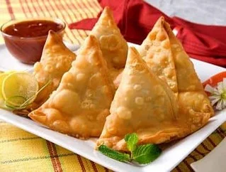 A plate of golden-brown samosas with a side of dipping sauce, garnished with a slice of lime and mint leaves.