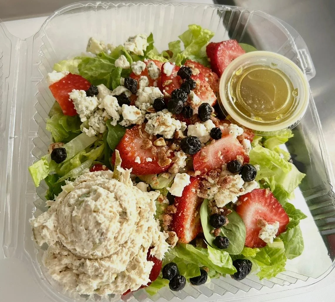 A fresh salad featuring lettuce, strawberries, blueberries, goat cheese, and a portion of creamy chicken salad, served with a side of dressing.