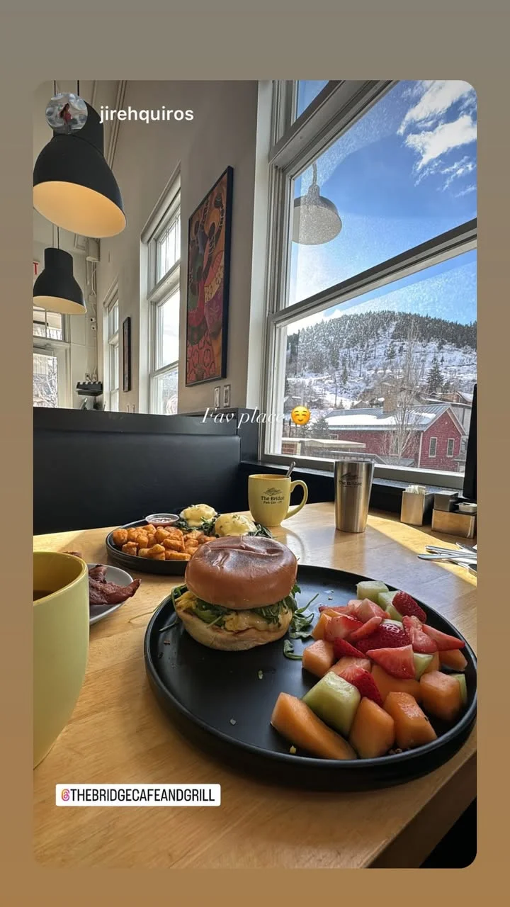 A cozy indoor dining setup featuring a delicious meal, including a sandwich and a fruit platter, with a scenic snowy view outside.