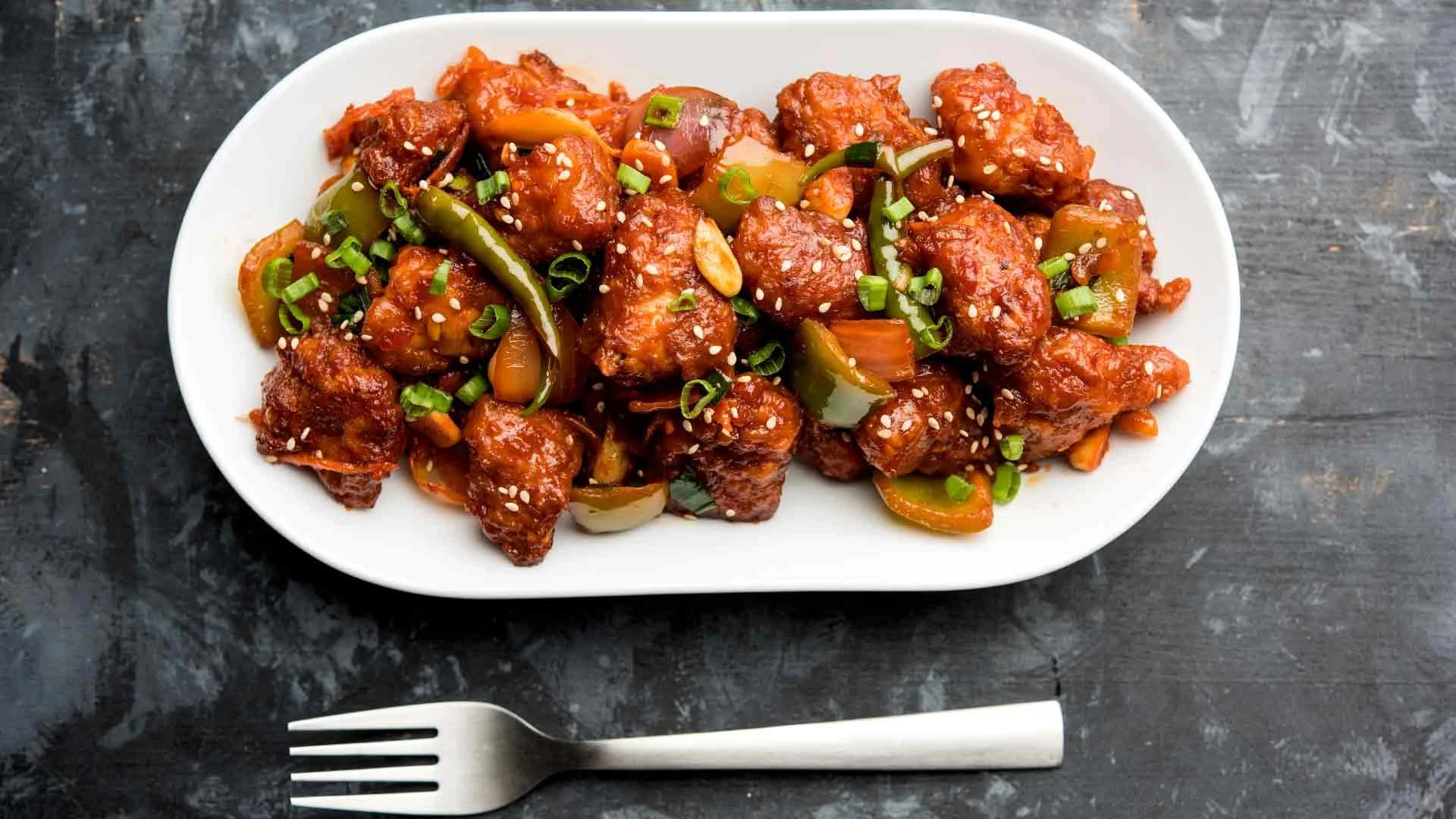 A dish of crispy, breaded pieces of chicken or tofu tossed in a rich, spicy sauce, garnished with green onions and sesame seeds. The dish includes slices of bell peppers.
