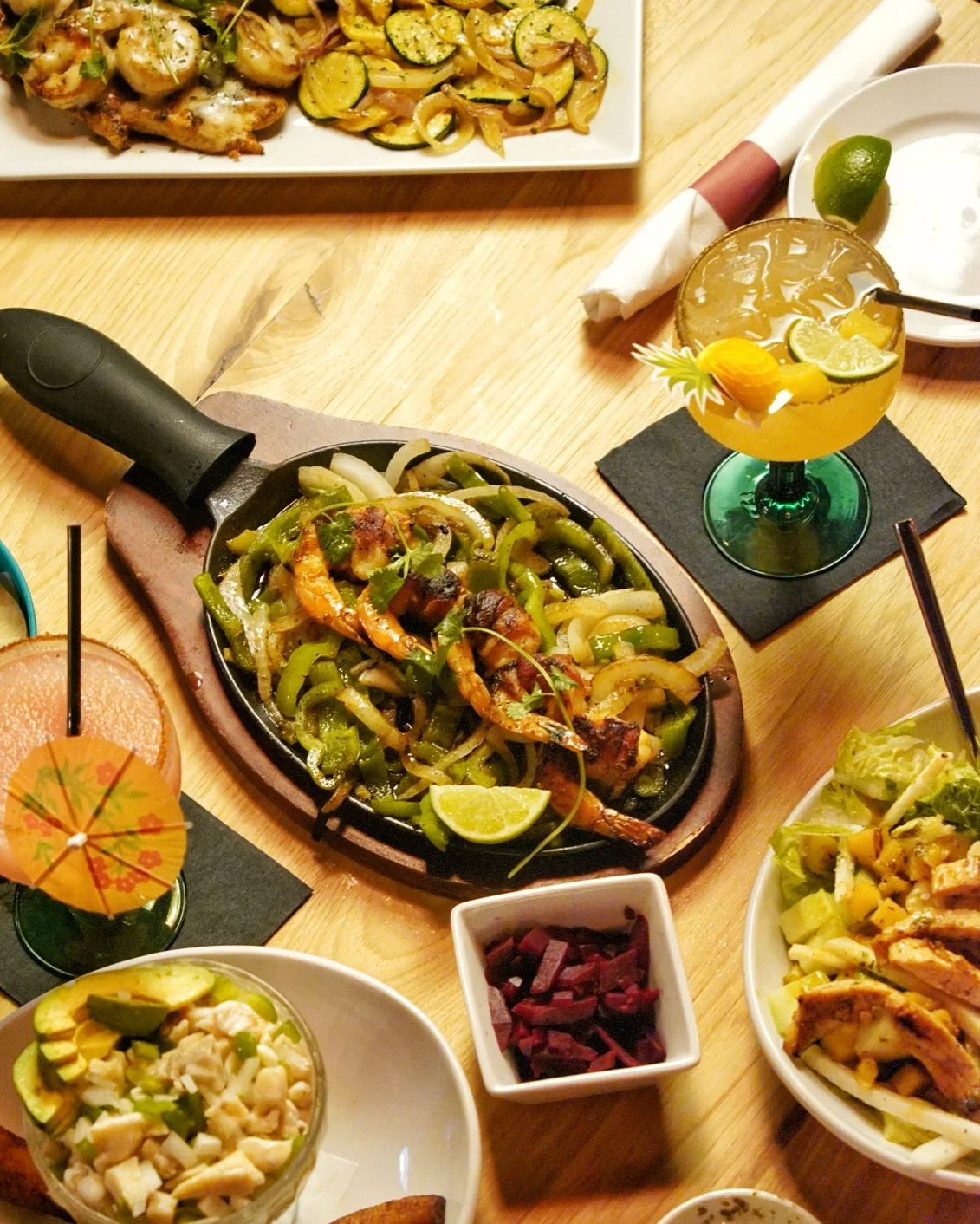 A sizzling plate of grilled shrimp and vegetables, including colorful bell peppers and onions, garnished with lime. Alongside, there are various side dishes and a vibrant cocktail.