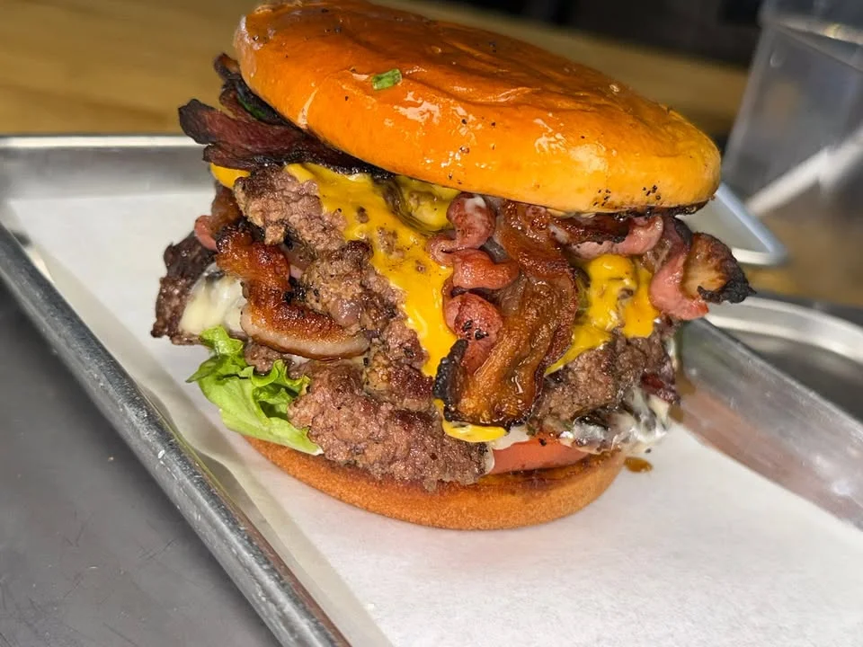 A large, stacked burger featuring layers of beef, cheese, and bacon, topped with lettuce and tomato on a shiny bun.