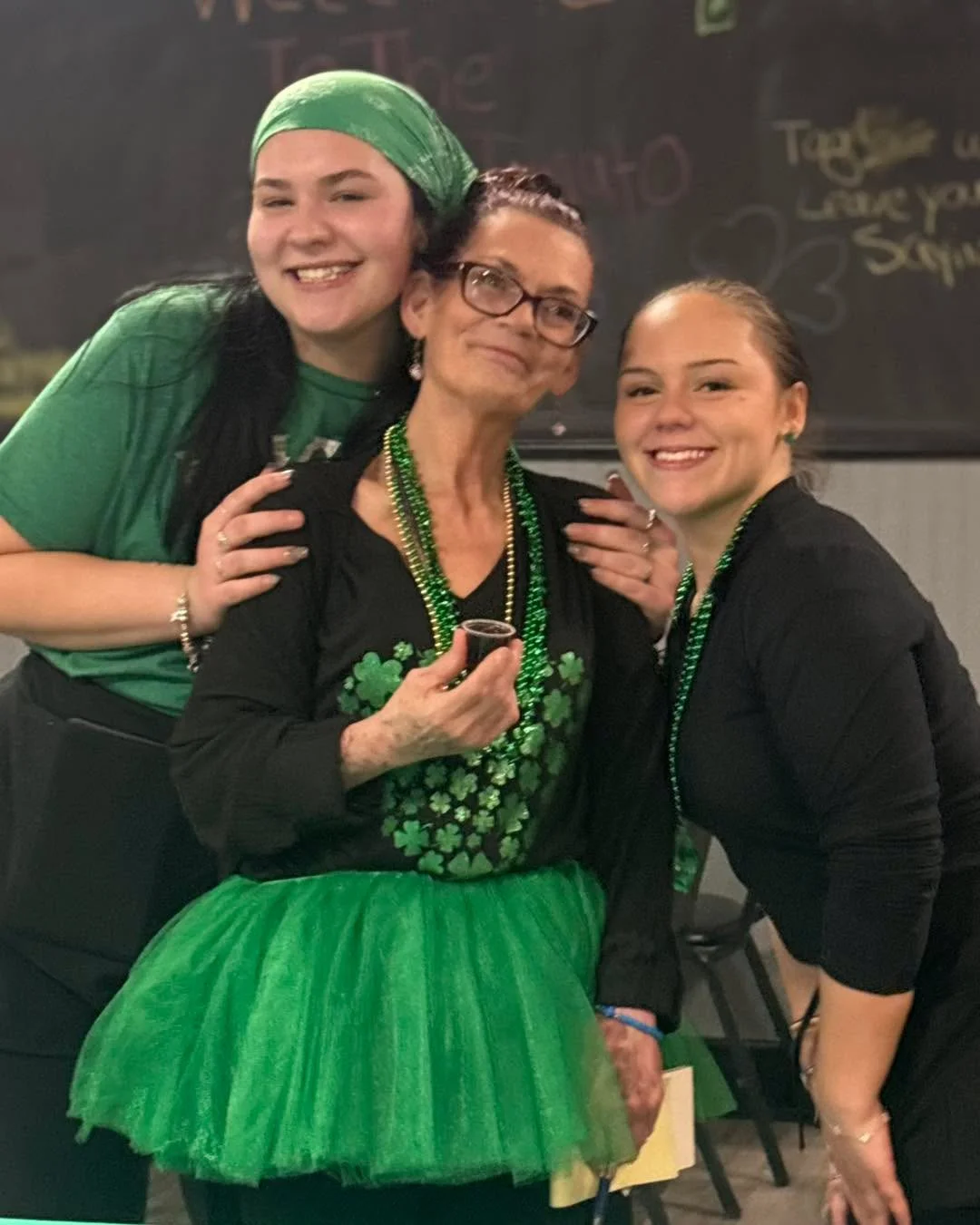 Three individuals in festive green attire posing together, celebrating with smiles. The backdrop suggests a lively dining or celebration scene.