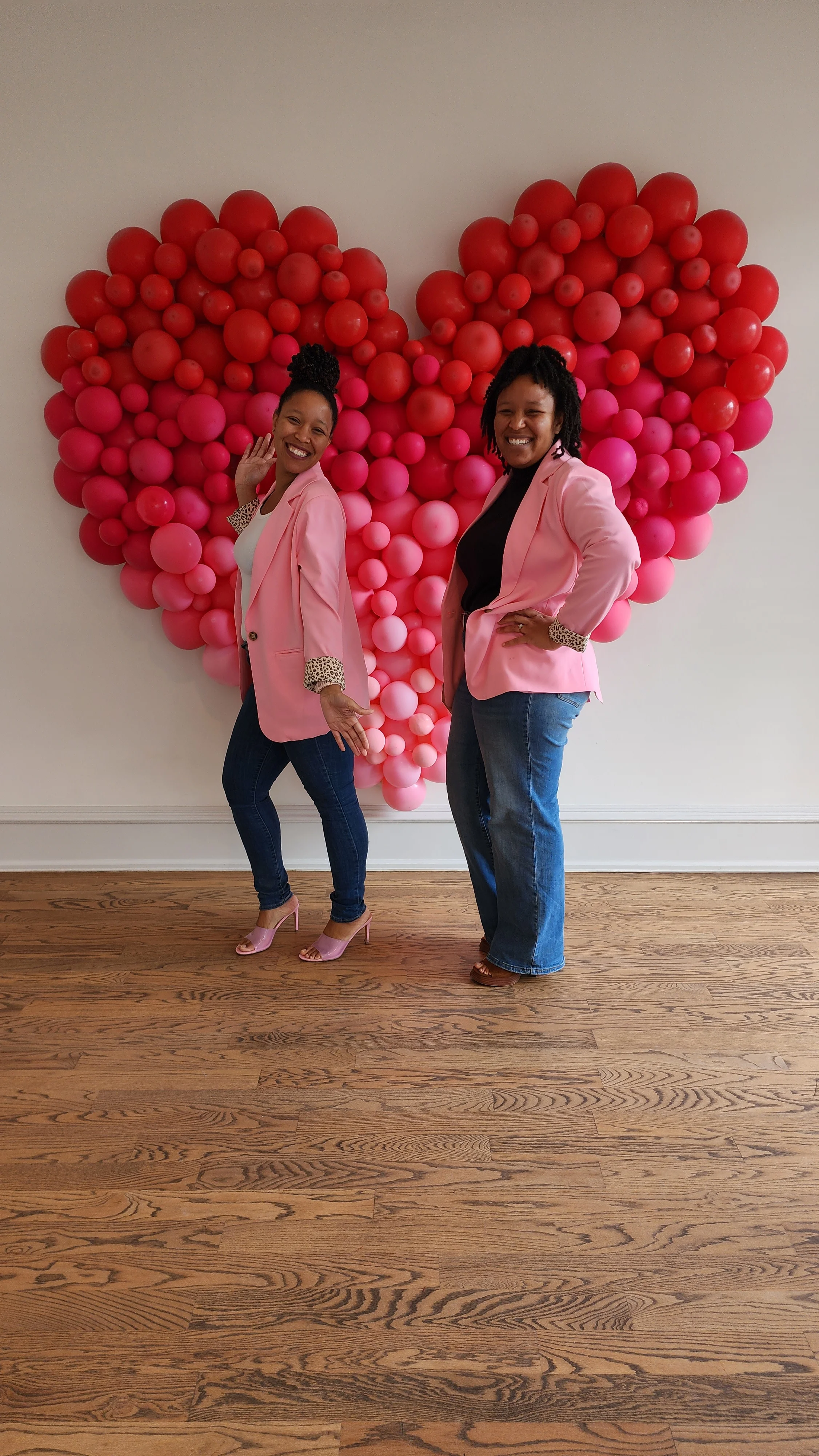Two women posing in pink blazers against a heart-shaped balloon backdrop