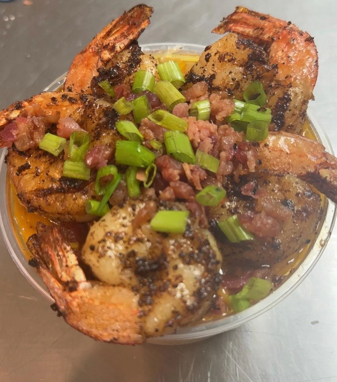 A serving of cooked shrimp topped with chopped green onions and bits of bacon, presented in a clear container.