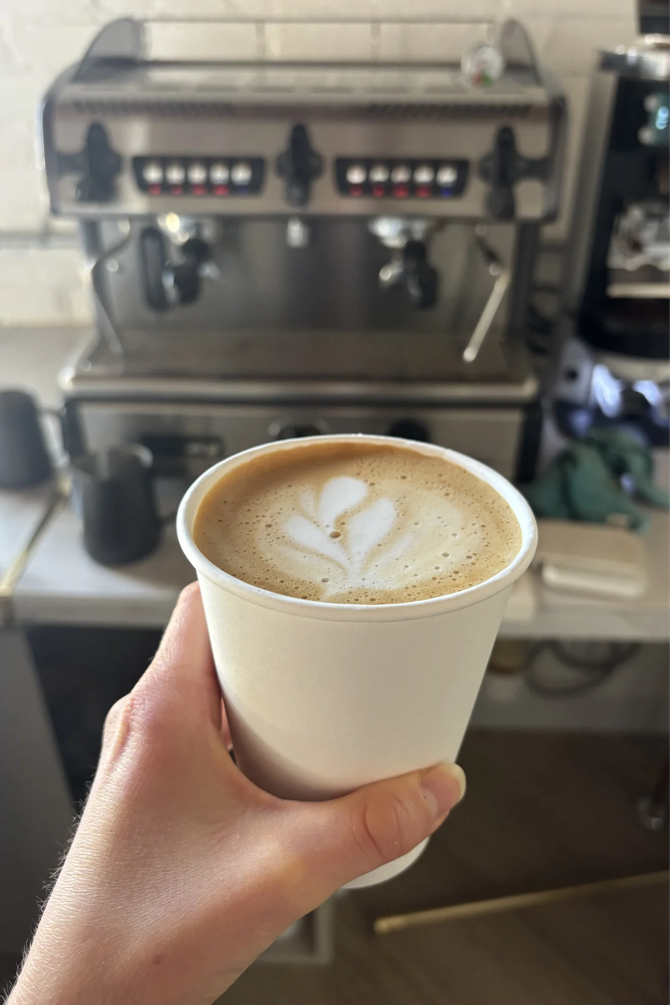 A hand holding a latte with latte art in a white cup, coffee machine in background.