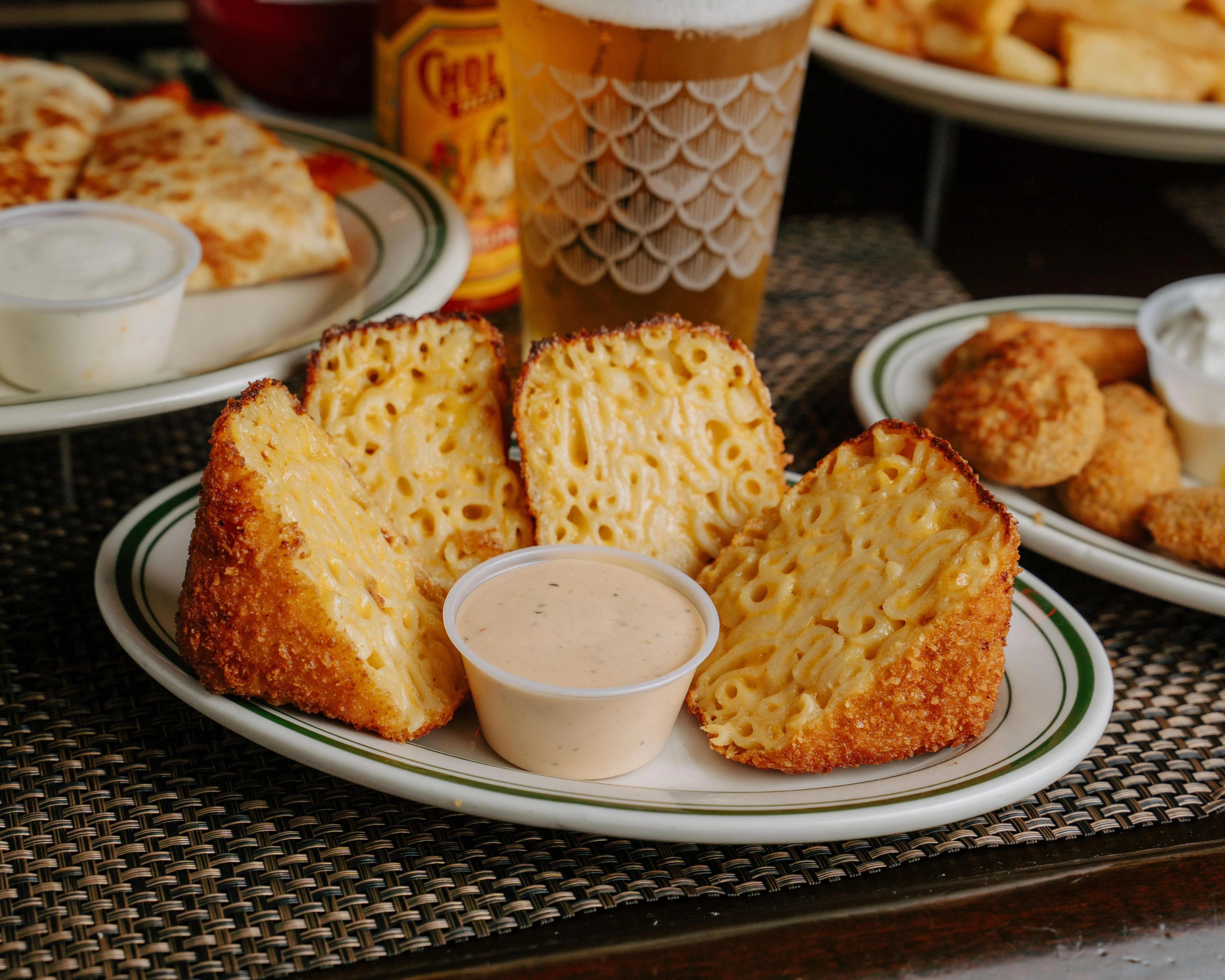 Fried macaroni and cheese wedges served with a dipping sauce on a plate. The wedges are golden brown and crispy, revealing a creamy macaroni filling.