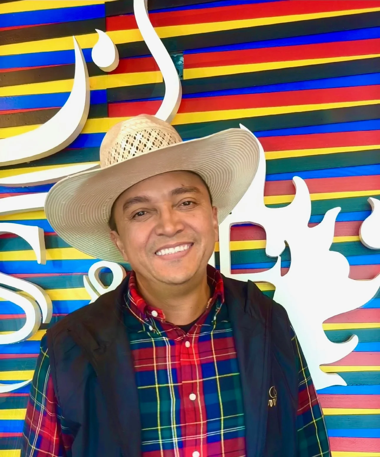 Smiling man in a plaid shirt and cowboy hat at a colorful restaurant backdrop