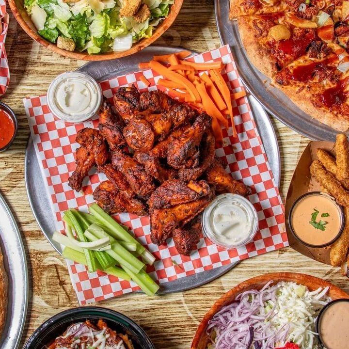 Overhead view of a tray of crispy chicken wings on red checkered paper, served with celery sticks, carrot sticks, and dipping sauces, with pizza and salad partially visible around the edges.