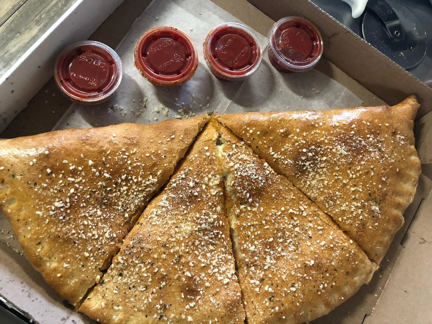 A large calzone sprinkled with garlic and herbs, cut into slices, accompanied by four small containers of marinara sauce.