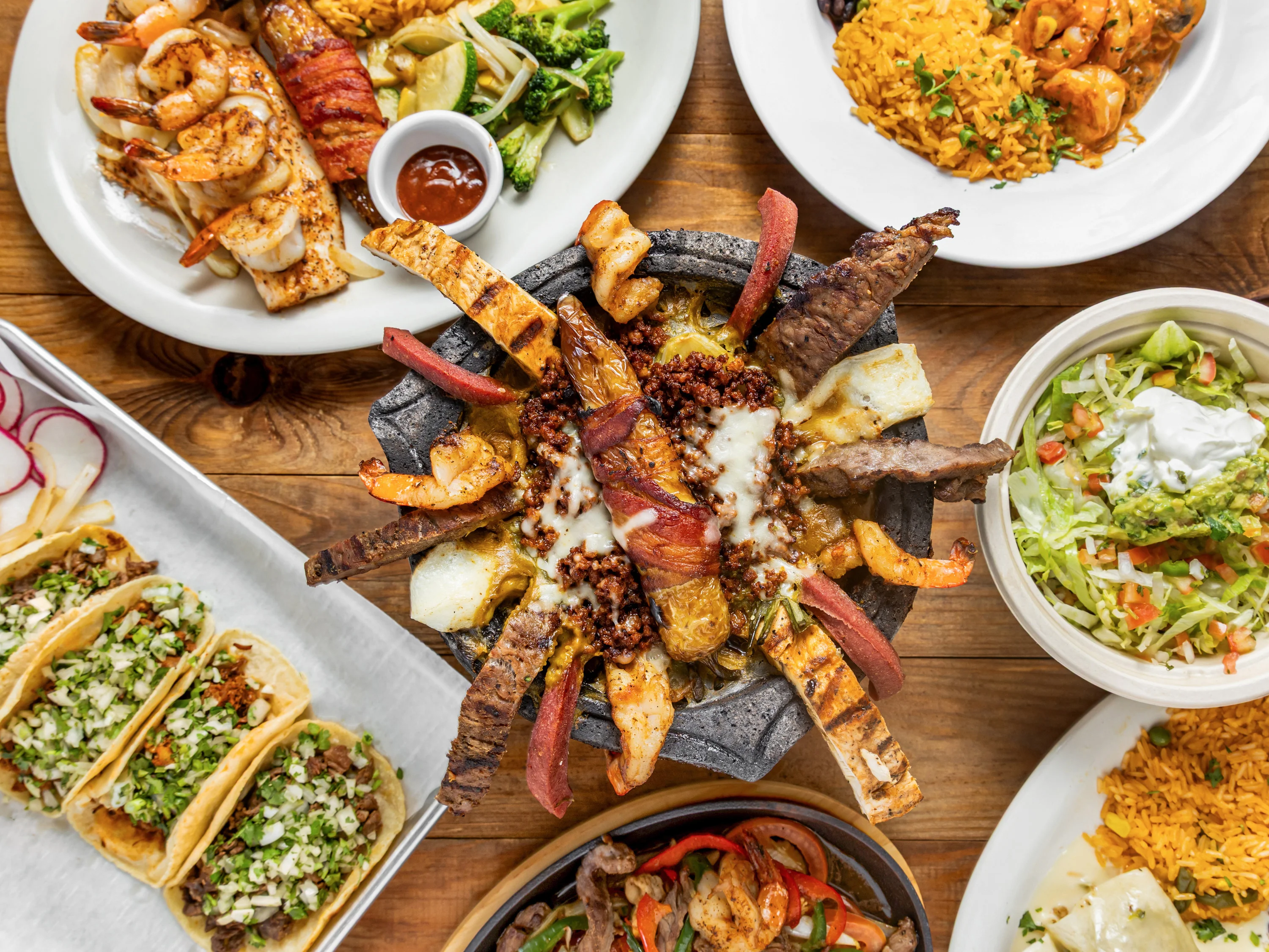 An assortment of Mexican dishes including grilled shrimp, tacos, and a sizzling meat platter, all beautifully arranged on a wooden table.