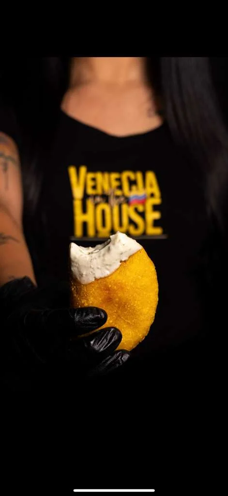 A person holding a golden, crispy food item with a creamy topping, showcasing a shirt that reads 'Venecia in the House'.