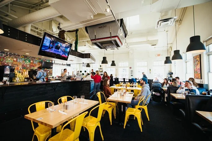 A busy restaurant interior showcasing yellow chairs and wooden tables. Diners are engaged with their meals and drinks, alongside a well-stocked bar.