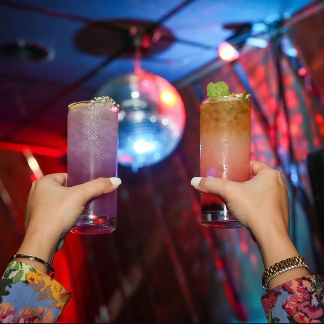 Two colorful cocktails are raised in celebration, with a disco ball reflecting light in the background. One drink is purple and the other, a gradient from amber to pink, garnished with a mint leaf.