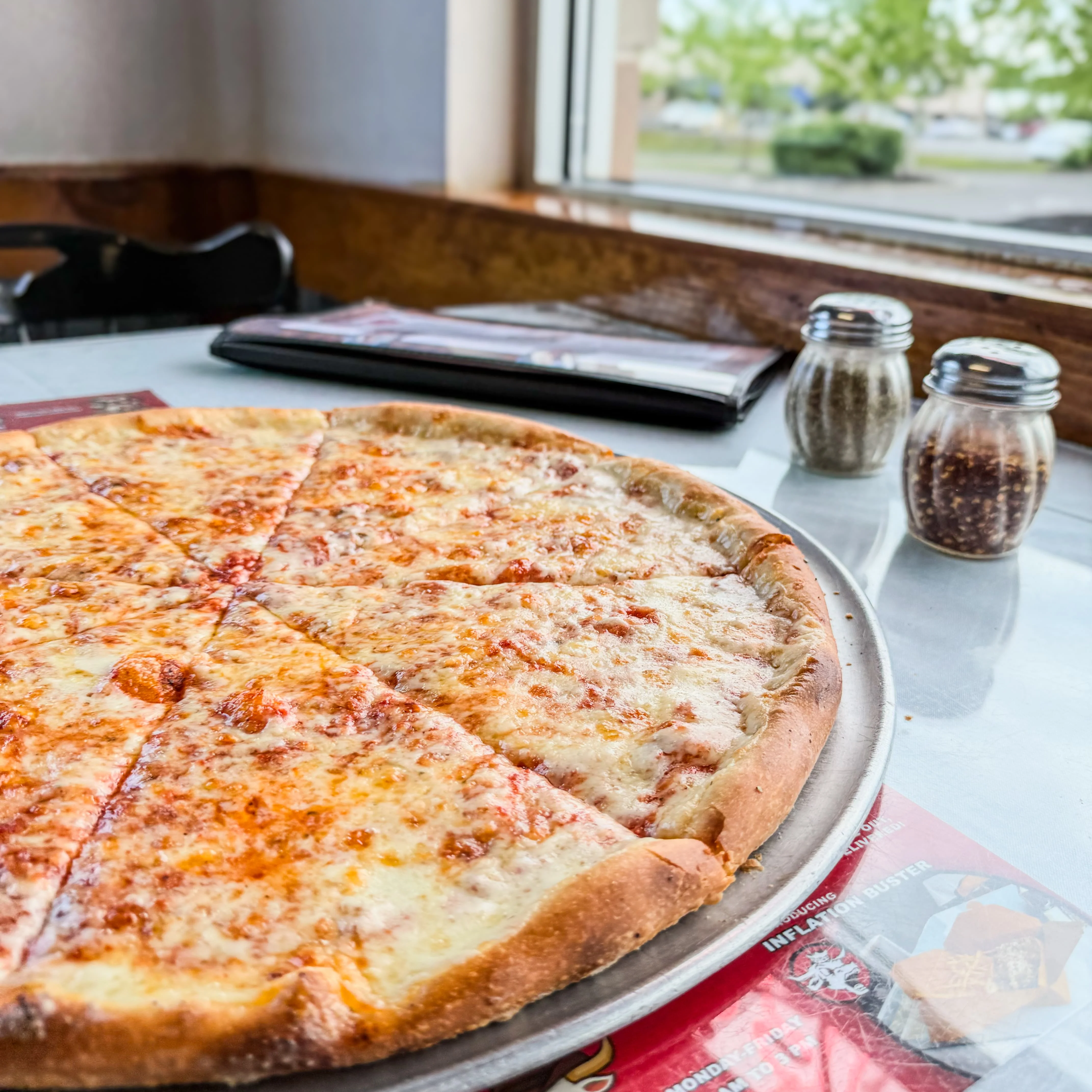  Full cheese pizza on silver tray near a window.